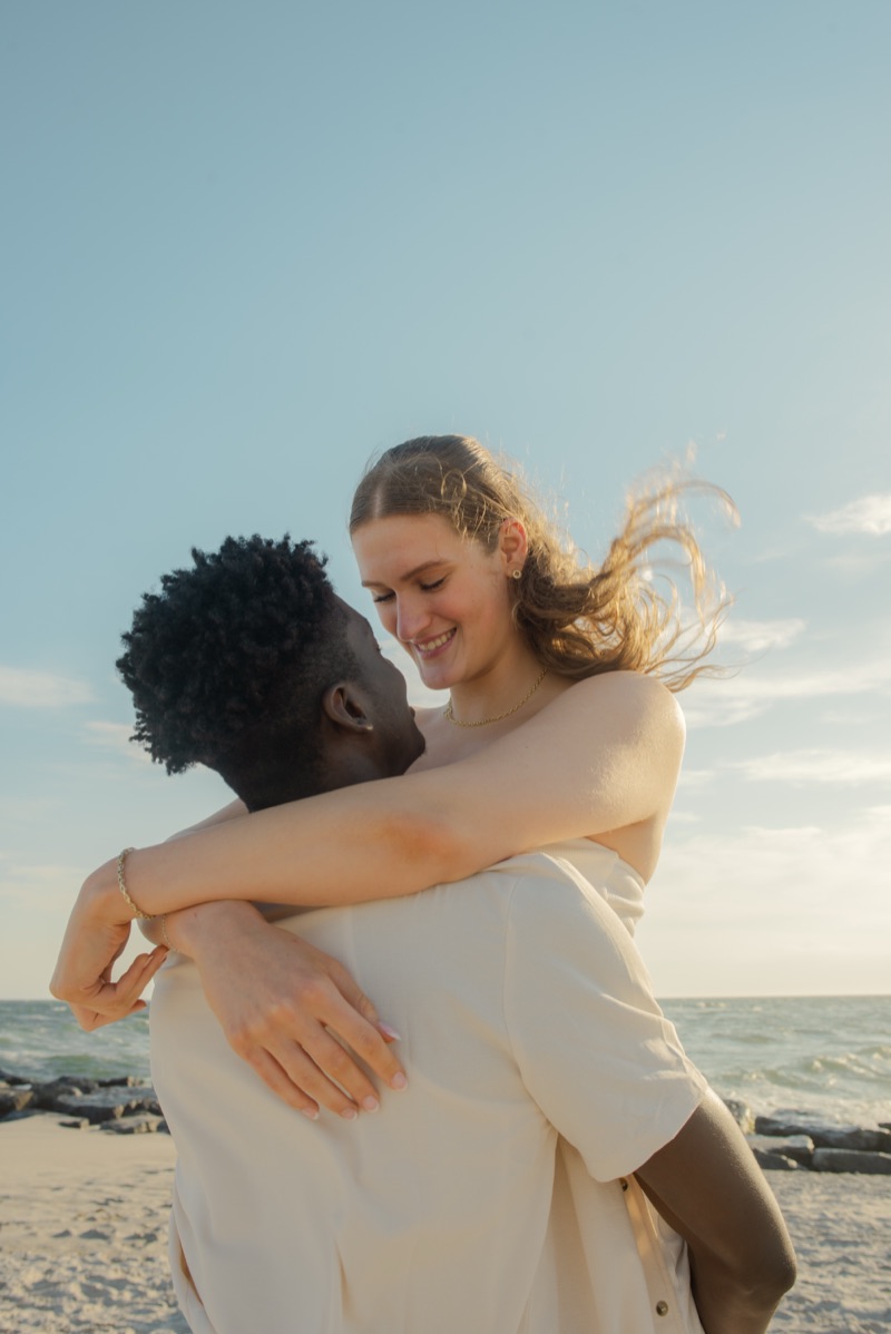 Backlit woman laughing with her arms around her partner’s shoulders on the beach at golden hour