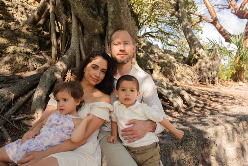 Parents in cream outfits seated among the roots of a large banyan tree with their two children on their laps