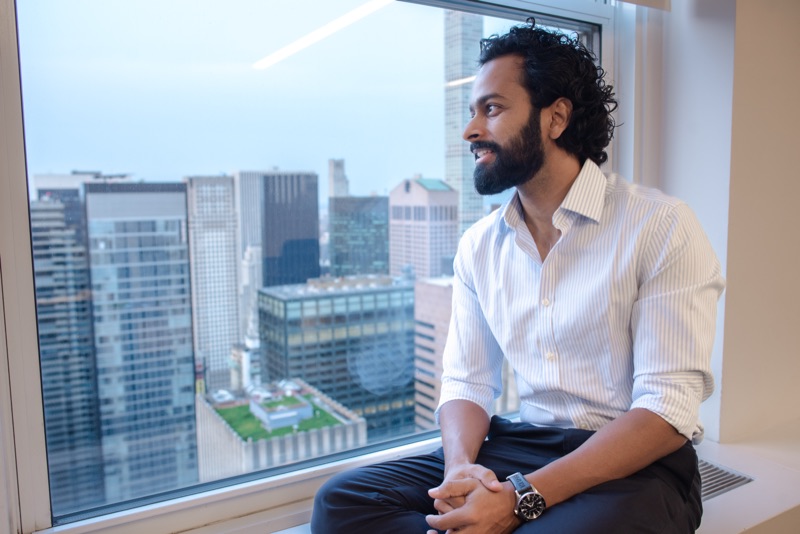 Man in a striped dress shirt seated on an office windowsill, smiling with Manhattan skyscrapers behind him