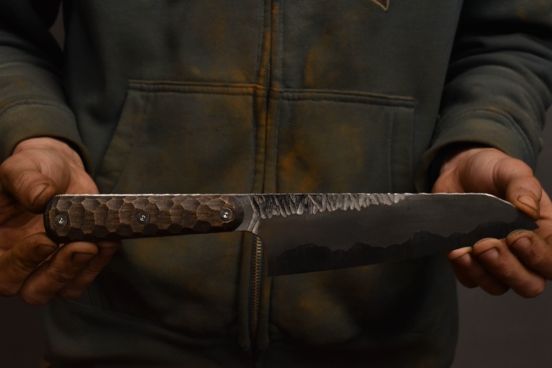 Hands holding a hand-forged kitchen knife with a carved wooden handle and a hammered blade