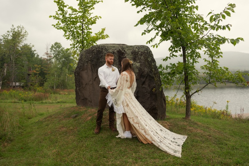 Groom holding the bride’s hand as her lace train spreads across the grass beside a mountain lake